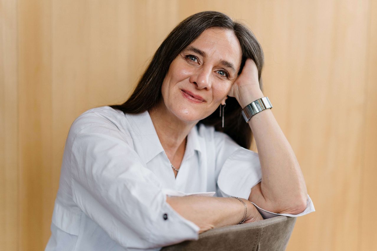 Eine Frau mit braunen Haaren, die ein weißes Hemd, eine Halskette und eine silberne Uhr trägt, ruht ihren Kopf auf ihrer Hand, während sie lächelt.