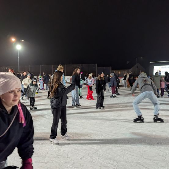 Viele Menschen skaten bei Nacht auf einer Eisbahn. Eine Person mit einer rosa Mütze lächelt in die Kamera. Eine Werbung auf einem Bildschirm ist sichtbar.