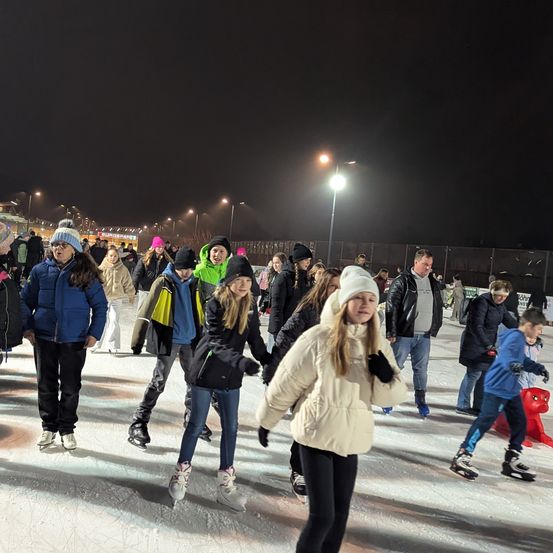 In der Nacht skatet eine Gruppe von Menschen, einschließlich Erwachsener und Kinder, auf einer Eisbahn im Freien. Der Himmel ist dunkel, und Straßenlaternen sind in der Ferne zu sehen.
