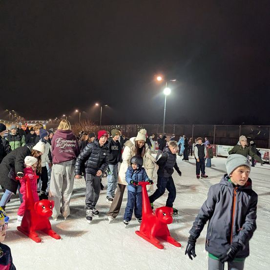 Bei Nacht genießen Menschen jeden Alters das Eislaufen auf einer Eisbahn. Einige nutzen rote Schlitten für zusätzlichen Spaß. Im Hintergrund befinden sich Straßenlaternen und ein Zaun.