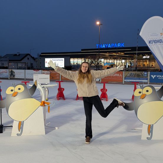 Eine Frau schlittert nachts vor einem Gebäude mit dem Schild 'THAYAPARK'. Sie trägt einen Pullover, eine Brille und Handschuhe. Zwei Pinguinstatuen stehen auf dem Eis.