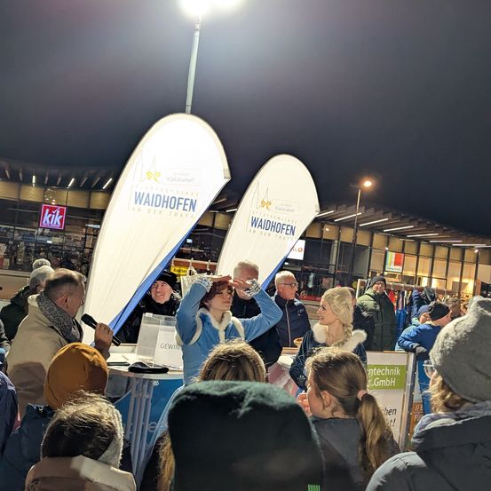 Ein Abendevent in einem Einkaufszentrum mit Bannern, die Waidhofen lesen. Menschen sind um einen Tisch versammelt, an dem eine Frau ein Mikrofon hält. Im Hintergrund ist ein Schild mit rotem Hintergrund zu sehen.