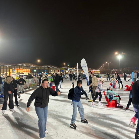 Eine Eisbahn im Freien bei Nacht mit vielen Schlittschuhläufern. Einige tragen Mützen und Handschuhe. Straßenlaternen beleuchten das Gebiet. In der Ferne sind Gebäude und ein Banner zu sehen. Pinguine sitzen auf dem Eis.