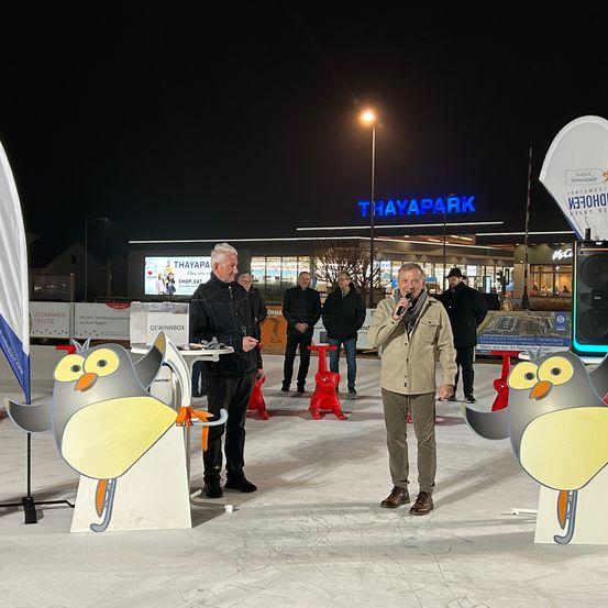 Ein Mann spricht in ein Mikrofon vor einer Pinguinstatue. Ein anderer Mann steht in der Nähe. Mehrere Leute beobachten im Hintergrund.