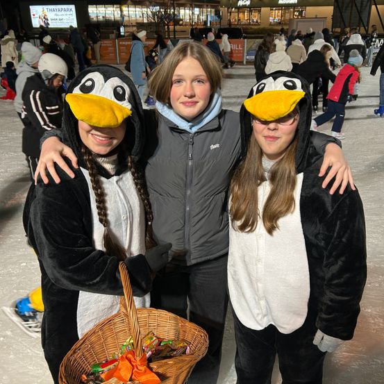 Drei Mädchen in Pinguinkostümen posieren für ein Foto auf einer Eisbahn, eine von ihnen hält einen Korb voller Süßigkeiten. Sie stehen zusammen und lächeln in die Kamera. Im Hintergrund sind mehrere Menschen in Winterkleidung, die auf dem Eis Schlittschuh laufen.