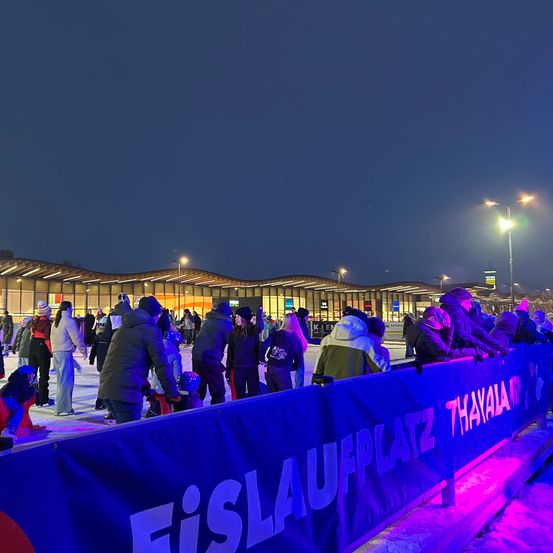 Eine Eislaufbahn bei Nacht, Leute skaten, einige sitzen in Stühlen, ein Banner steht 'Eislaufplatz That.' Gebäude und Straßenlaternen sind sichtbar.