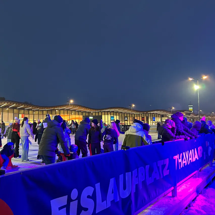 Eine Eislaufbahn bei Nacht, Leute skaten, einige sitzen in Stühlen, ein Banner steht 'Eislaufplatz That.' Gebäude und Straßenlaternen sind sichtbar.