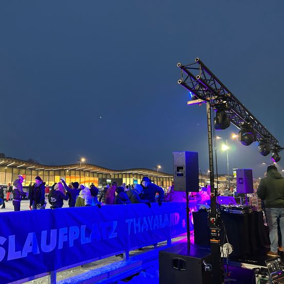Eine Nachtveranstaltung mit Menschen, die auf einer Eisbahn Schlittschuh laufen, umgeben von einem blauen Banner und Lautsprechern.