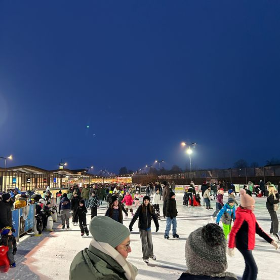 Eine Eisbahn im Freien mit vielen Menschen, die Schlittschuh laufen und herumstehen. Es gibt Straßenlaternen und ein Gebäude im Hintergrund. Es ist Nacht.