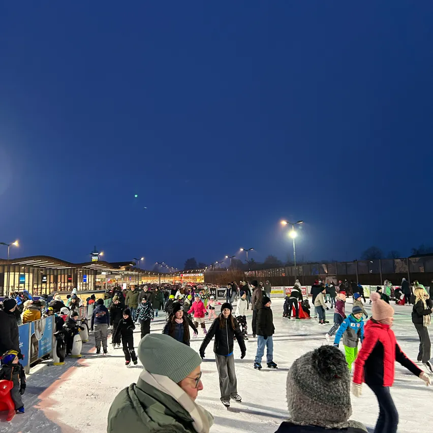Eine Eisbahn im Freien mit vielen Menschen, die Schlittschuh laufen und herumstehen. Es gibt Straßenlaternen und ein Gebäude im Hintergrund. Es ist Nacht.