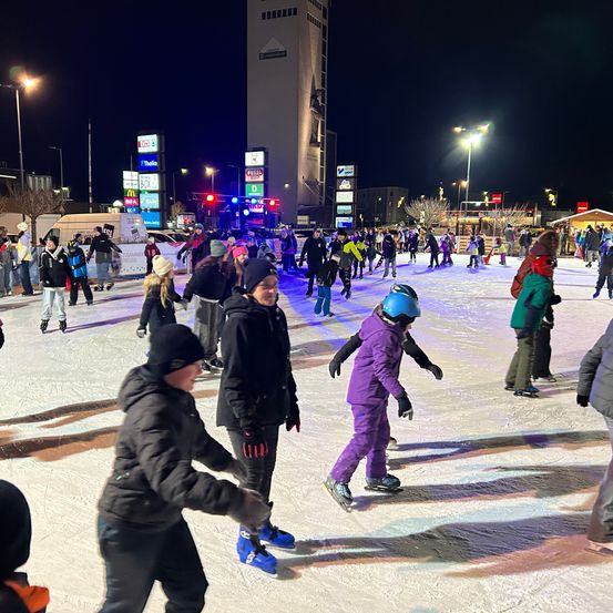 Viele Menschen skaten bei Nacht auf einer Freiluft-Eisbahn. Im Hintergrund steht ein hoher Turm. Es gibt viele Straßenlaternen und Schilder.