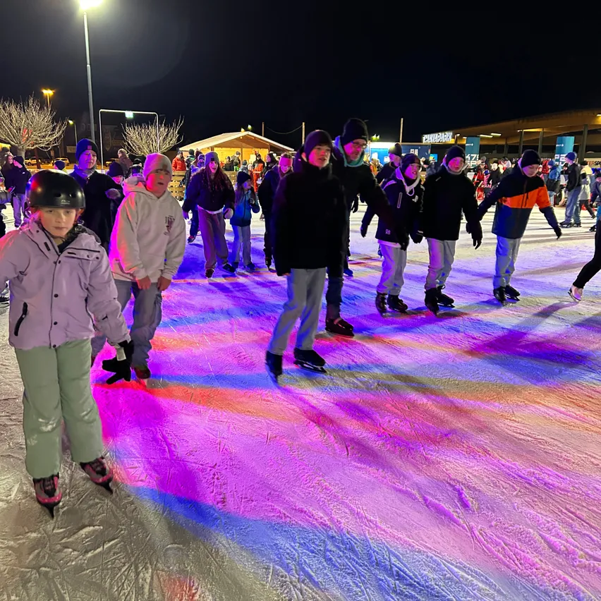 In der Nacht skaten mehrere Menschen auf einer beleuchteten Eisbahn mit einer farbenfrohen Lichtshow auf dem Boden.