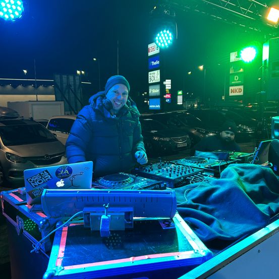 Ein DJ steht bei einer DJ-Konsole mit Neonlichtern und einem geparkten Auto vor einem McDonald's-Schild bei Nacht.