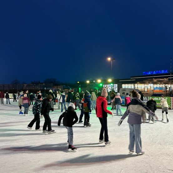 Eine Eisbahn bei Nacht, mehrere Menschen sind beim Eislaufen. Ein Schild im Hintergrund lautet Thaypark.