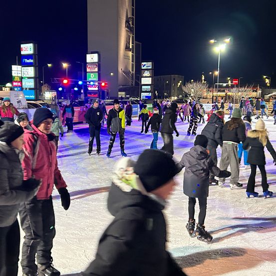 Bei Nacht skatet eine Menschenmenge auf einer Eisbahn im Freien. Sie tragen Winterkleidung und Schlittschuhe. Werbung und Straßenlaternen umgeben die Eisbahn.