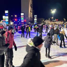 Bei Nacht skatet eine Menschenmenge auf einer Eisbahn im Freien. Sie tragen Winterkleidung und Schlittschuhe. Werbung und Straßenlaternen umgeben die Eisbahn.