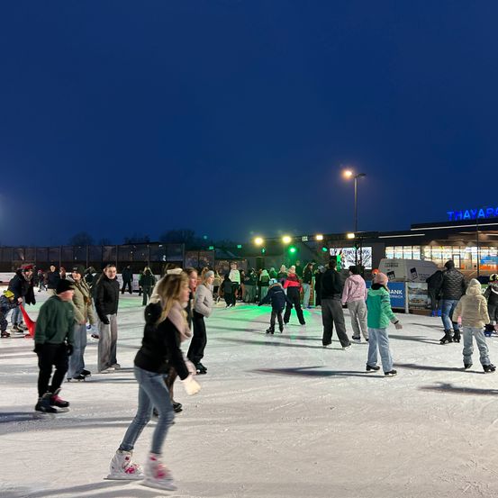 Menschen Schlittschuh laufen bei Nacht auf einer Freiluftbahn mit einem Gebäude im Hintergrund und Straßenlaternen.
