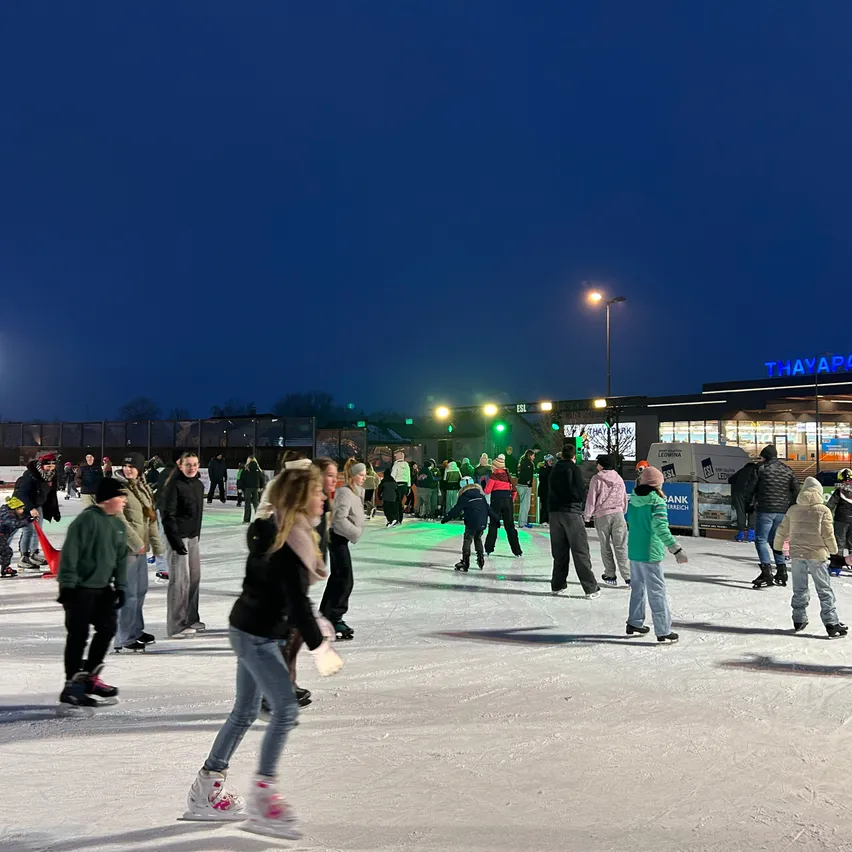 Menschen Schlittschuh laufen bei Nacht auf einer Freiluftbahn mit einem Gebäude im Hintergrund und Straßenlaternen.