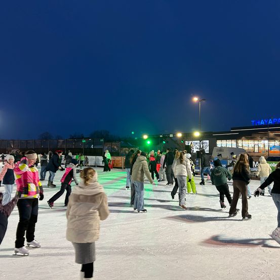 Menschen skaten bei Nacht auf einer Eislaufbahn im Freien, beleuchtet von Straßenlaternen. Im Hintergrund ist ein Gebäude mit einem Schild mit der Aufschrift THAYAPA zu sehen.