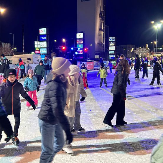Eine Gruppe von Menschen, die nachts auf einer Freiluft-Eisbahn Schlittschuh laufen. Sie tragen Winterkleidung und Schlittschuhe. Es gibt Werbung an den Straßenlaternen und Gebäuden.