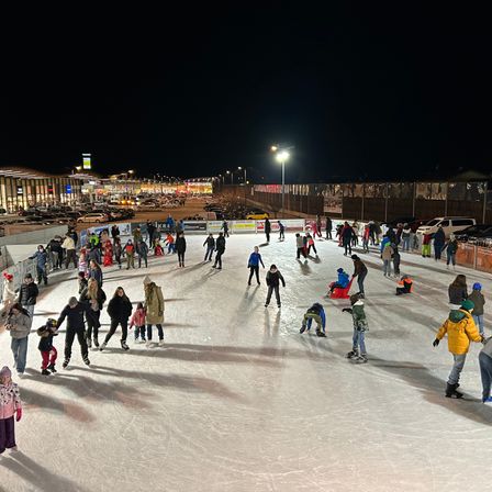 Eine große Gruppe von Menschen fährt bei Nacht auf einer Eisbahn im Freien Schlittschuh, mit mehreren geparkten Autos im Hintergrund.