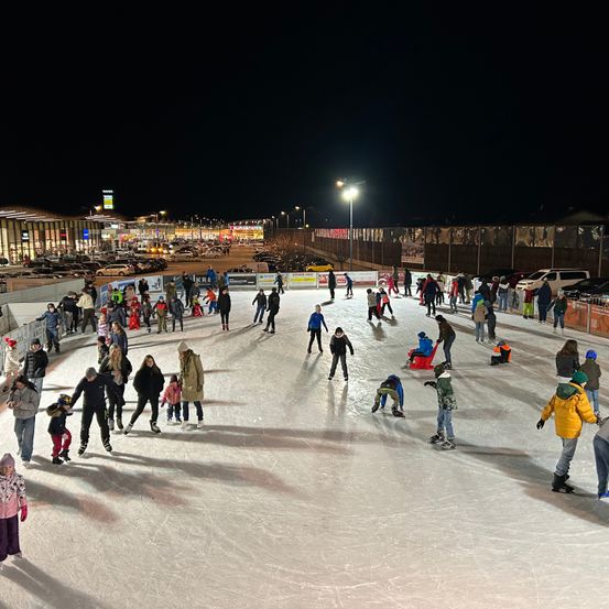 Eine große Gruppe von Menschen fährt bei Nacht auf einer Eisbahn im Freien Schlittschuh, mit mehreren geparkten Autos im Hintergrund.