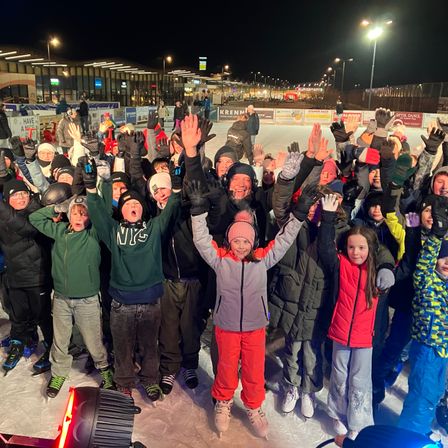 Eine Gruppe von Menschen steht bei Nacht auf einer Eisbahn, lächelt und hebt die Hände. Sie tragen alle Winterkleidung.
