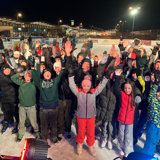 Eine Gruppe von Menschen steht bei Nacht auf einer Eisbahn, lächelt und hebt die Hände. Sie tragen alle Winterkleidung.