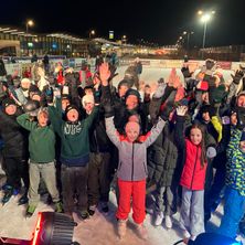 Eine Gruppe von Menschen steht bei Nacht auf einer Eisbahn, lächelt und hebt die Hände. Sie tragen alle Winterkleidung.