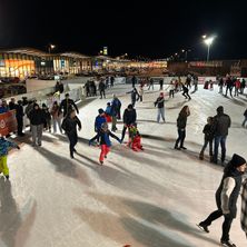 Eine Eisbahn im Freien ist bei Nacht beleuchtet. Viele Menschen, einschließlich Kinder, eislaufen. Straßenlaternen und ein Schild sind im Hintergrund zu sehen.