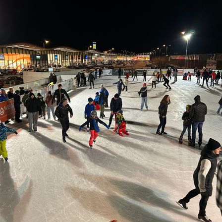 Eine Eisbahn im Freien ist bei Nacht beleuchtet. Viele Menschen, einschließlich Kinder, eislaufen. Straßenlaternen und ein Schild sind im Hintergrund zu sehen.
