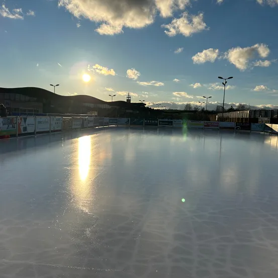 Eine Eisbahn im Freien unter einem klaren Himmel bei Sonnenuntergang. Das Eis ist glatt, und es gibt Werbebanner rund um die Bahn. Straßenlaternen säumen die Seiten.