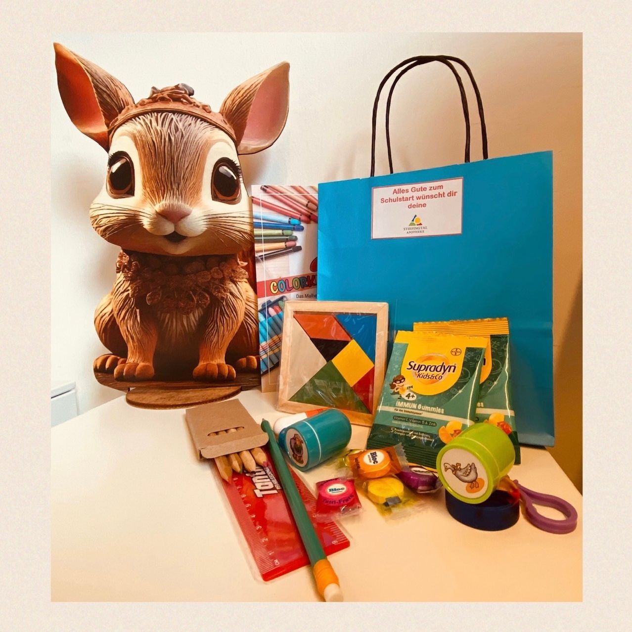 A table displays a bunny figurine, a blue bag with a label, a box of colored pencils, a ruler, a green pen, and various other items.