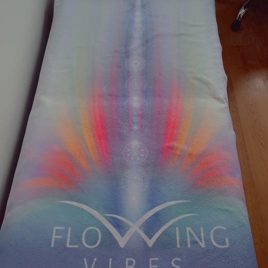Eine farbenfrohe Yogamatte mit einem abstrakten, bunten Design. Das Wort 'FLOWING' ist prominent in Weiß angebracht und die Matte liegt auf einem Holzboden.