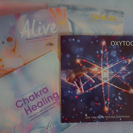 Vier Bücher liegen auf einer weißen Oberfläche. 'Alive' ist links mit einem Babygesicht. 'Avalon' ist in der Mitte mit einem Hirsch. 'Oxytocin' ist rechts mit Händen, die ein leuchtendes Atom berühren. 'Chakra Healing' ist unter 'Alive' mit einer Hand und leuchtenden Linien.