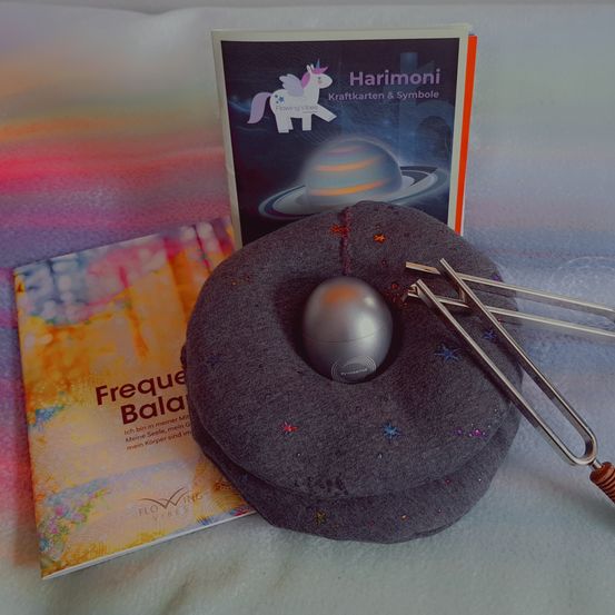 Ein Kissen mit einem Gong und einem Buch mit dem Titel 'Freque Bala'. Dahinter ein Buch mit einem Einhorn und dem Titel 'Harimoni'. Der Hintergrund ist farbenfroh.