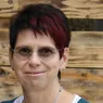 Eine Frau mit kurzen schwarzen und roten Haaren lächelt und trägt eine Brille. Sie hat eine Halskette und Ohrringe. Hinter ihr befindet sich eine Holzwand.