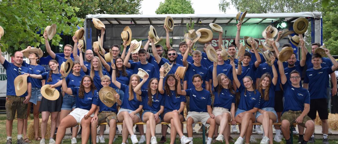 Eine Gruppe von Menschen in blauen T-Shirts posiert für ein Foto, einige halten Hüte in der Hand, auf Bänken in einem Park mit Bäumen im Hintergrund.