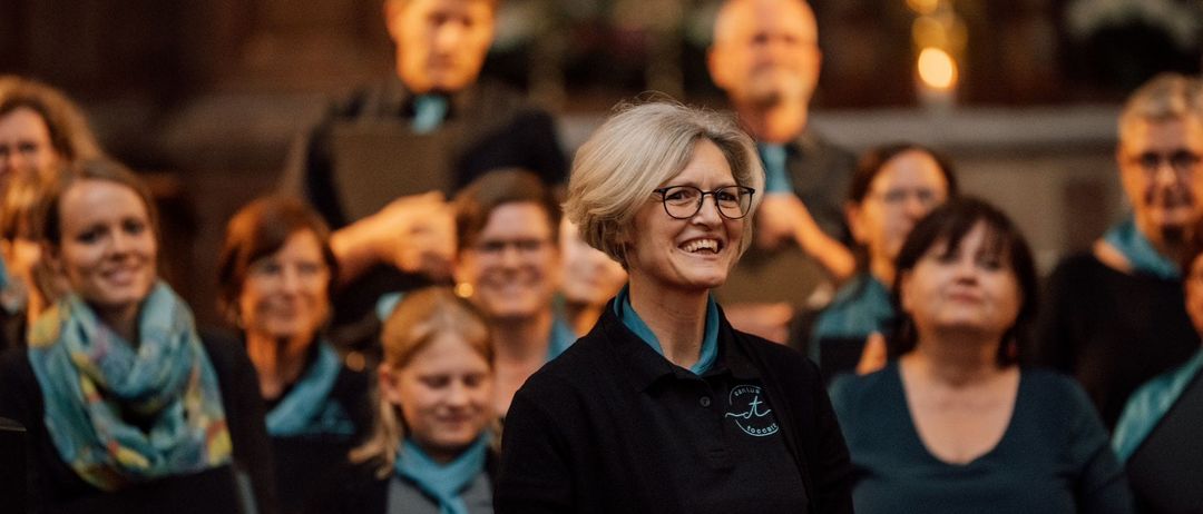 Eine Frau mit Brille und einem schwarzen Poloshirt lächelt vor einem verschwommenen Publikum, wahrscheinlich bei einer musikalischen Veranstaltung.