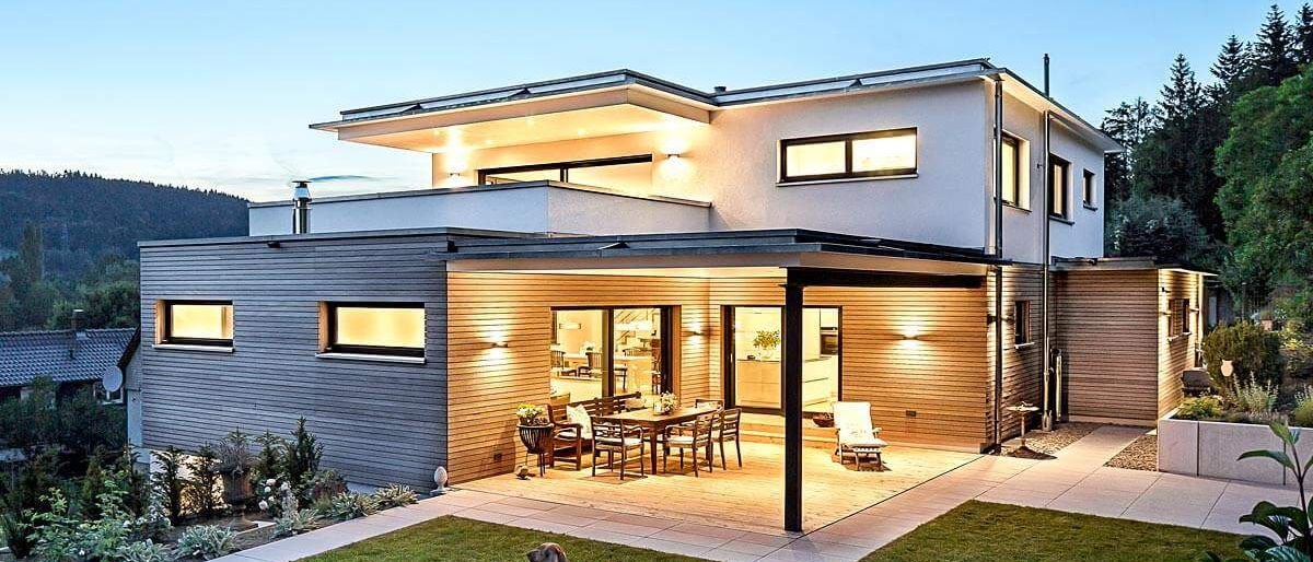 Modernes Hausaußenbereich bei Dämmerung mit einem Hund auf dem Rasen, mit überdachter Terrasse, Esstisch und offener Küche.