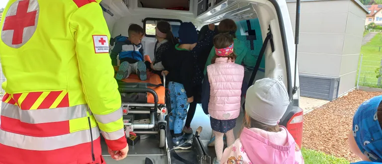 Ein Sanitäter kümmert sich um ein kleines Kind in einem Krankenwagen, während mehrere Zuschauer, darunter Kinder, zusehen.