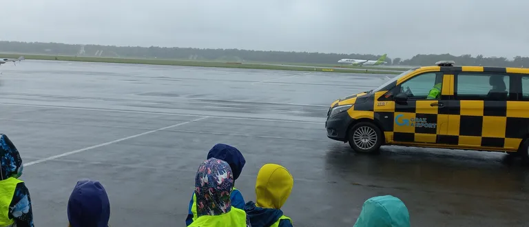 Kinder in Regenkleidung stehen auf einer nassen Startbahn und beobachten einen gelben und schwarzen Taxi, der vorbeifährt. Ein Flugzeug ist in der Ferne sichtbar.