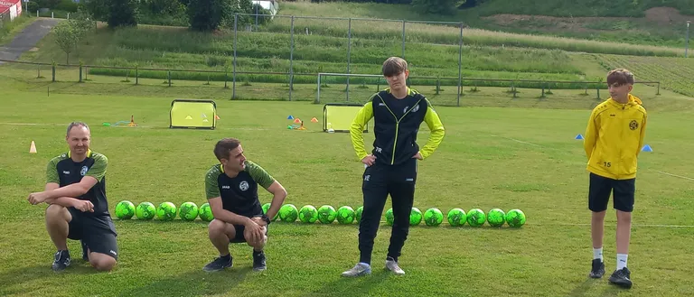 Drei Personen befinden sich auf einem Rasenfeld, möglicherweise für eine Fußballtrainingseinheit. Zwei knien, einer steht. Sie sind von Fußballbällen und Trainingshütchen umgeben. Im Hintergrund steht ein Haus und Bäume.