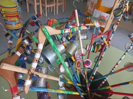 Eine bunte Ansammlung von Holz- und Metallobjekten hängt von einer zentralen Struktur, einschließlich verschiedener Dosen und Glocken. Der Ort scheint ein Innenraum zu sein, mit Holzstühlen und einem gelben Hintergrund.