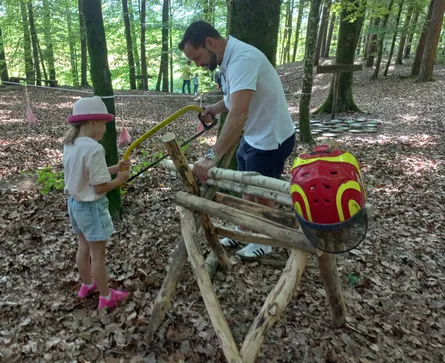 Ein Mann hilft einem kleinen Mädchen, einen Baumstamm mit einer Säge in einem Wald zu schneiden. Ein Helm liegt auf dem Baumstamm.