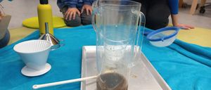 Ein durchsichtiger Krug mit brauner Flüssigkeit steht auf einem Tablett, umgeben von einem Trichter, einem Filter, einer gelben Flasche und einem Becher auf einem blauen Tuch. Zwei Personen sitzen hinter dem Tablett.