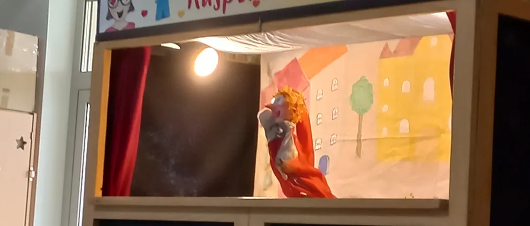 Eine Puppentheaterbühne mit einem farbenfrohen Hintergrund, der Gebäude und einen Baum zeigt. Eine Puppe mit blondem Haar ist auf der Bühne.
