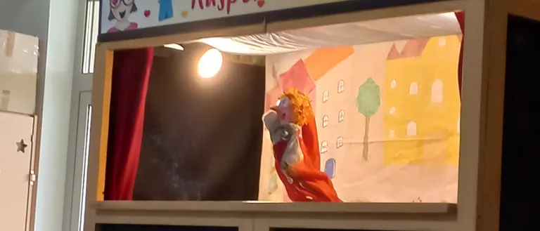 Eine Puppentheaterbühne mit einem farbenfrohen Hintergrund, der Gebäude und einen Baum zeigt. Eine Puppe mit blondem Haar ist auf der Bühne.
