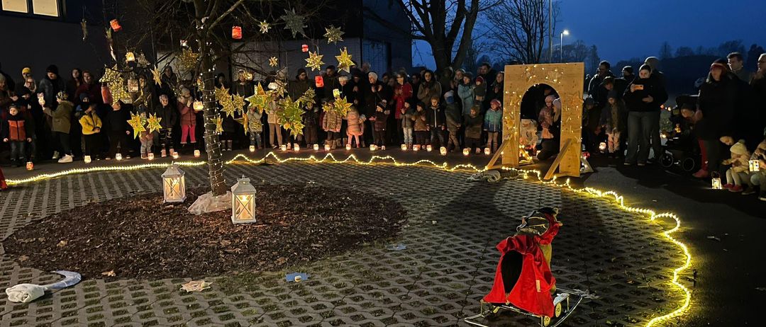 Eine Menschenmenge steht um einen dekorierten Baum herum und hält brennende Laternen in der Nacht. Ein Schlitten mit einem roten Mantel liegt auf dem Boden, umgeben von einem gelben Licht.