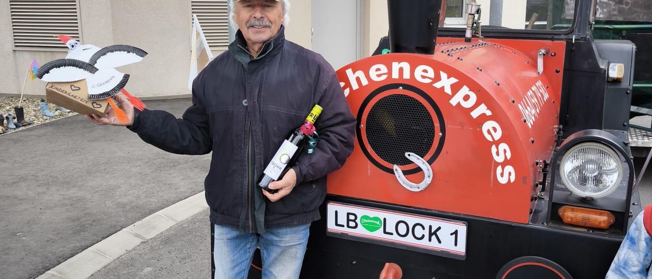 Ein älterer Mann steht neben einem roten Zug, hält eine Flasche Wein und einen Vogel. Der Zug hat ein Schild mit der Aufschrift 'chenexpress'.